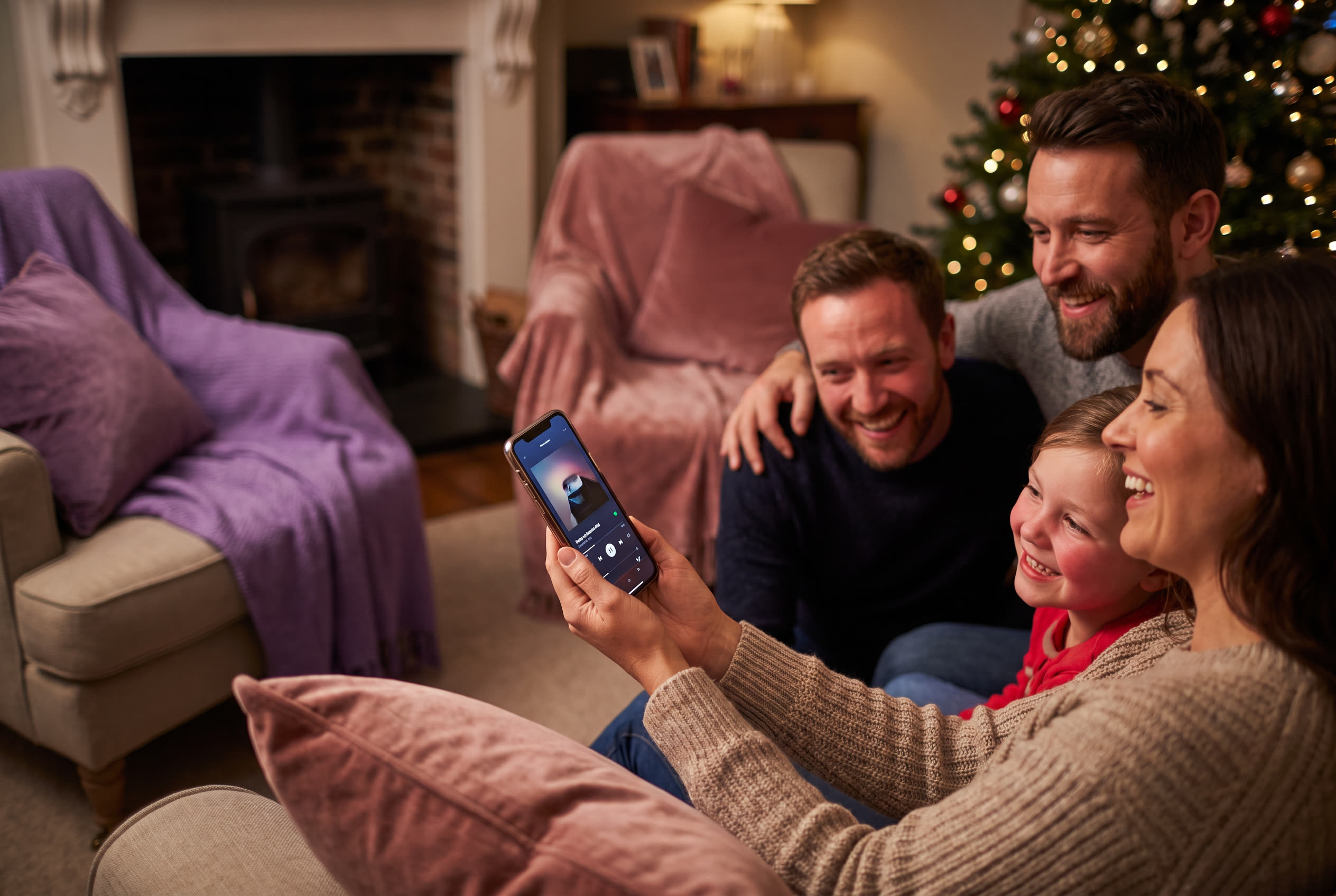 A family playing a song from a smartphone for their best friend during a christmas celebration, everyone listening with delighted smiles
